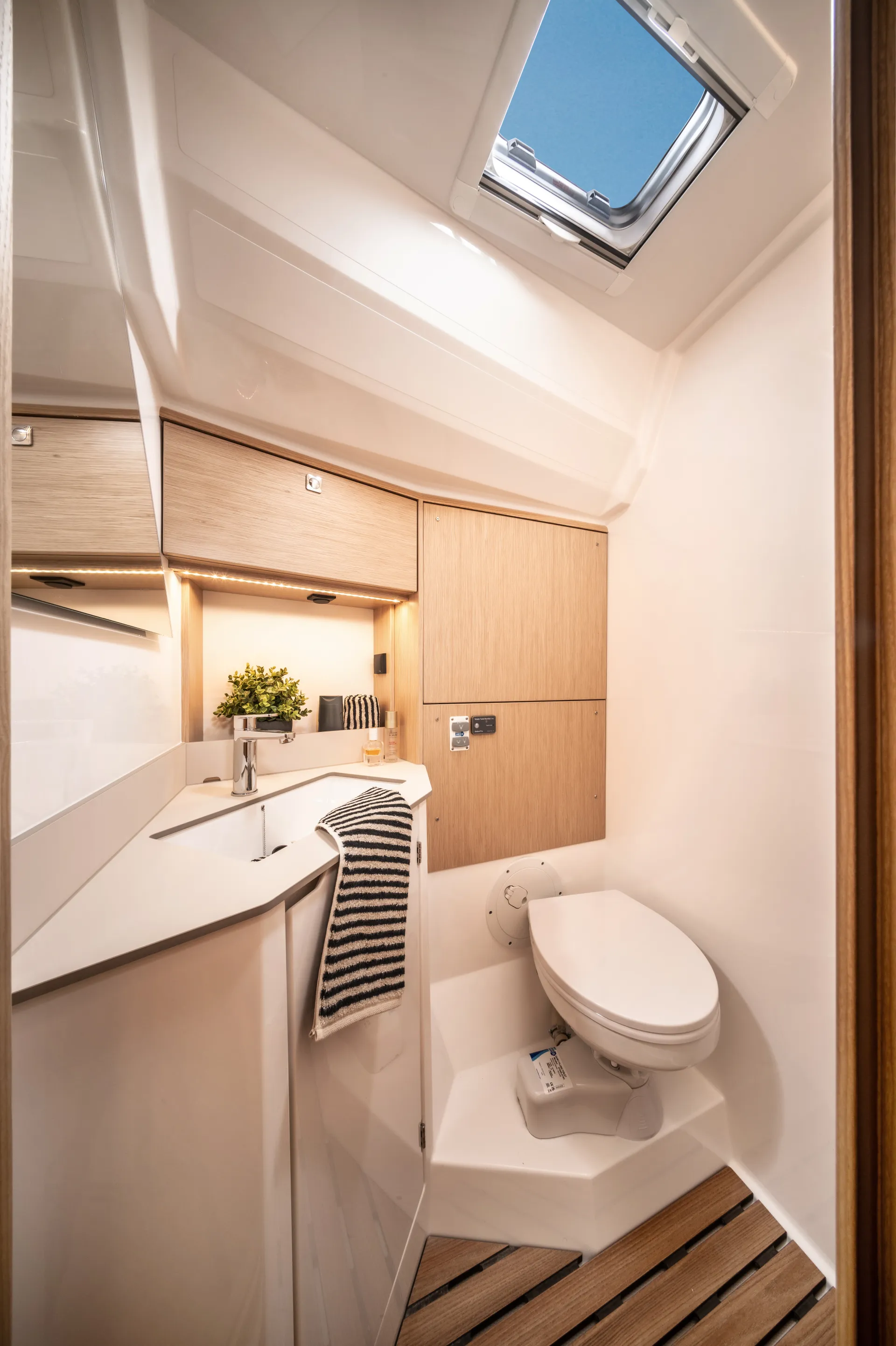 Modern boat bathroom with wood accents, a compact white basin, striped towel, shelves with toiletries, small potted plant, and a white toilet under a skylight. Natural light brightens the clean, organised space.