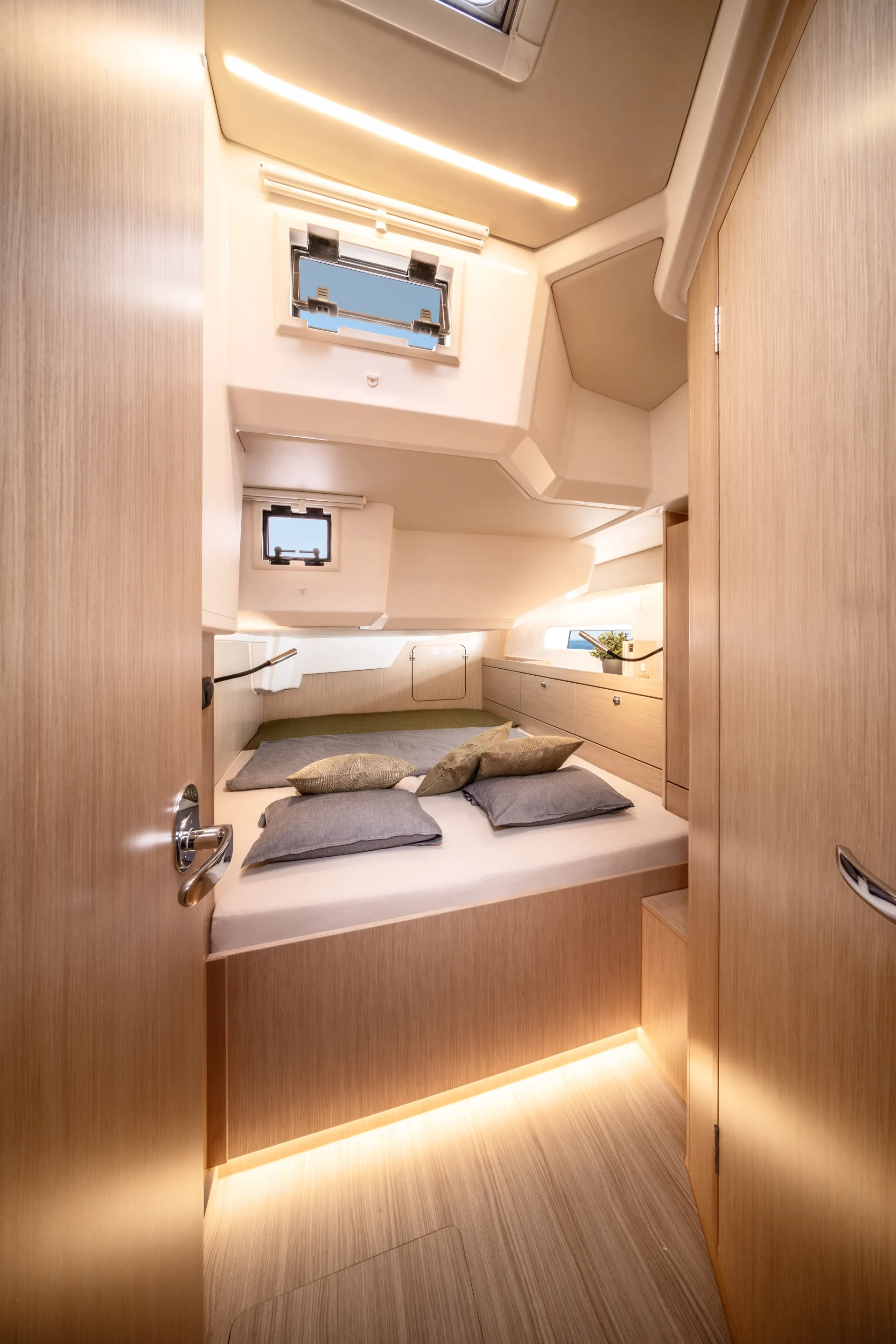 A cosy, modern boat cabin with wood panelling, a bed with grey and green pillows, two small windows, soft lighting, and built-in storage with a plant on a shelf.