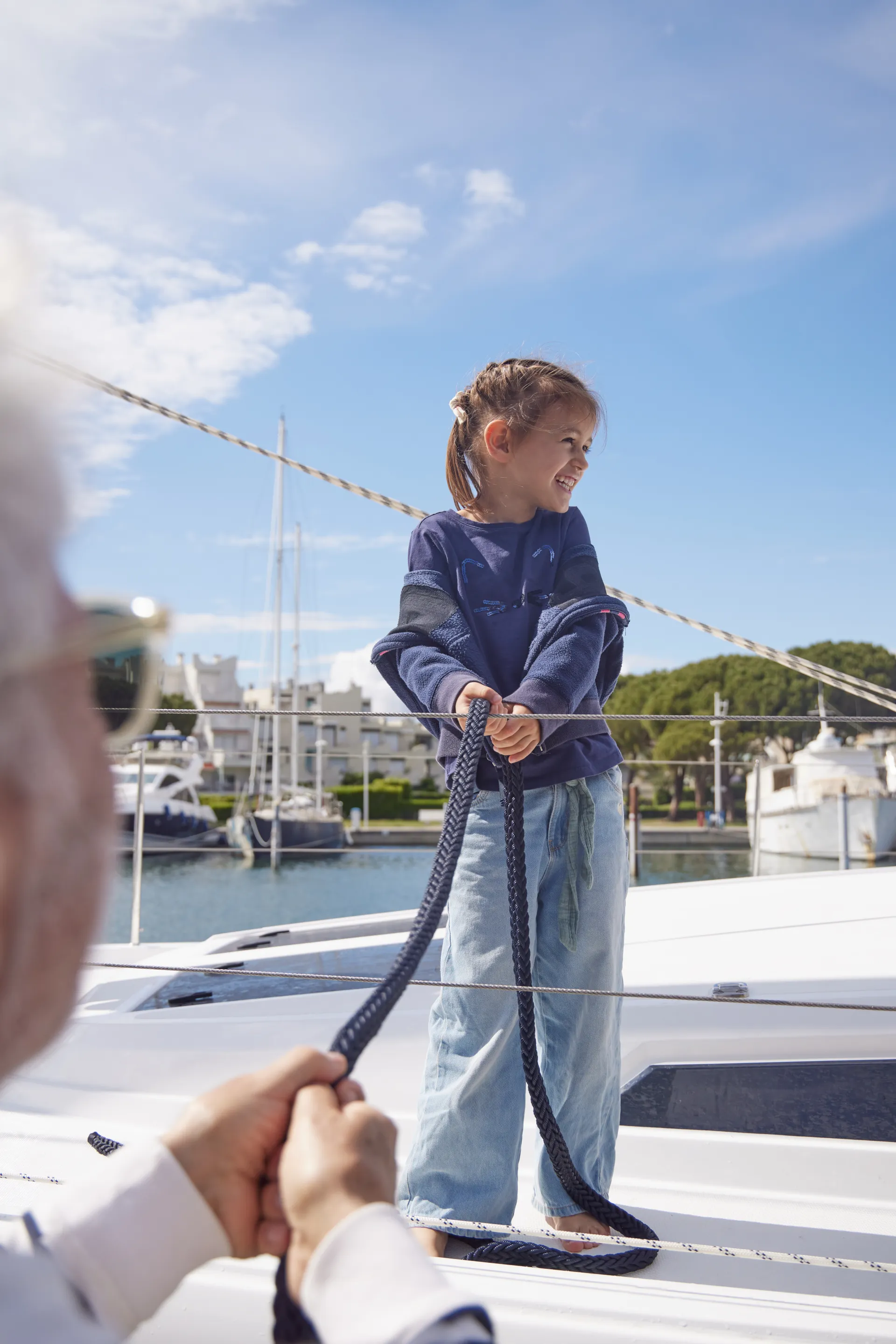 A smiling child stands on a boat holding a rope, with an adult assisting. Boats and a marina are visible in the background under a blue sky.