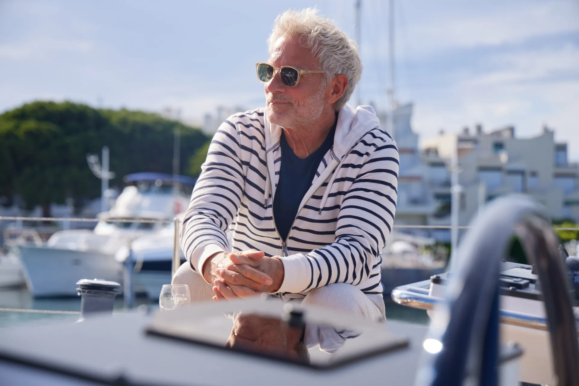 Older man with grey hair and sunglasses, wearing a striped hoodie, sits relaxed on a boat at a marina with yachts and buildings in the background on a sunny day.