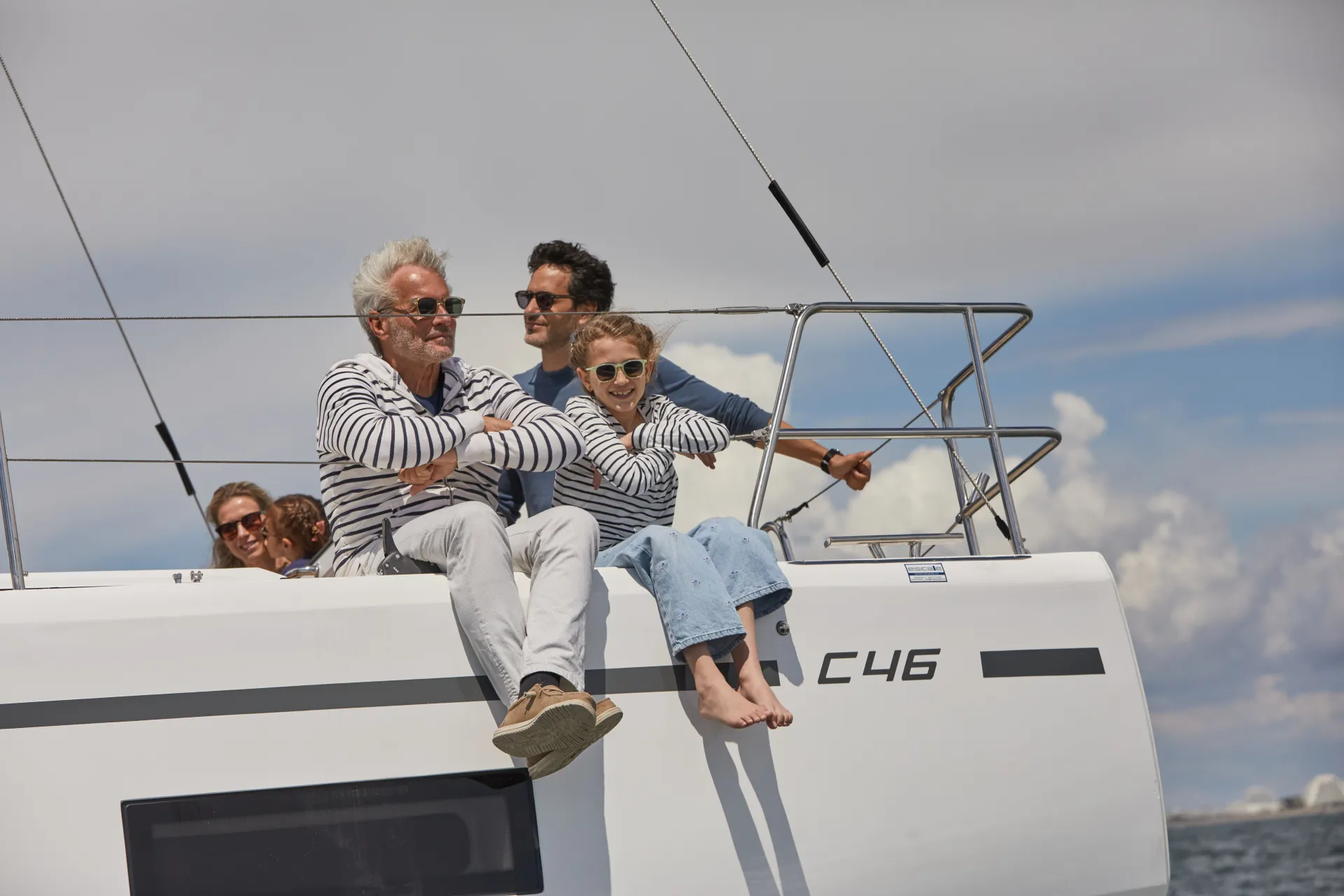 Four people relax on the deck of a sailing boat under a partly cloudy sky. Two are seated at the edge in matching striped shirts, while two others sit behind them, smiling and enjoying the view.